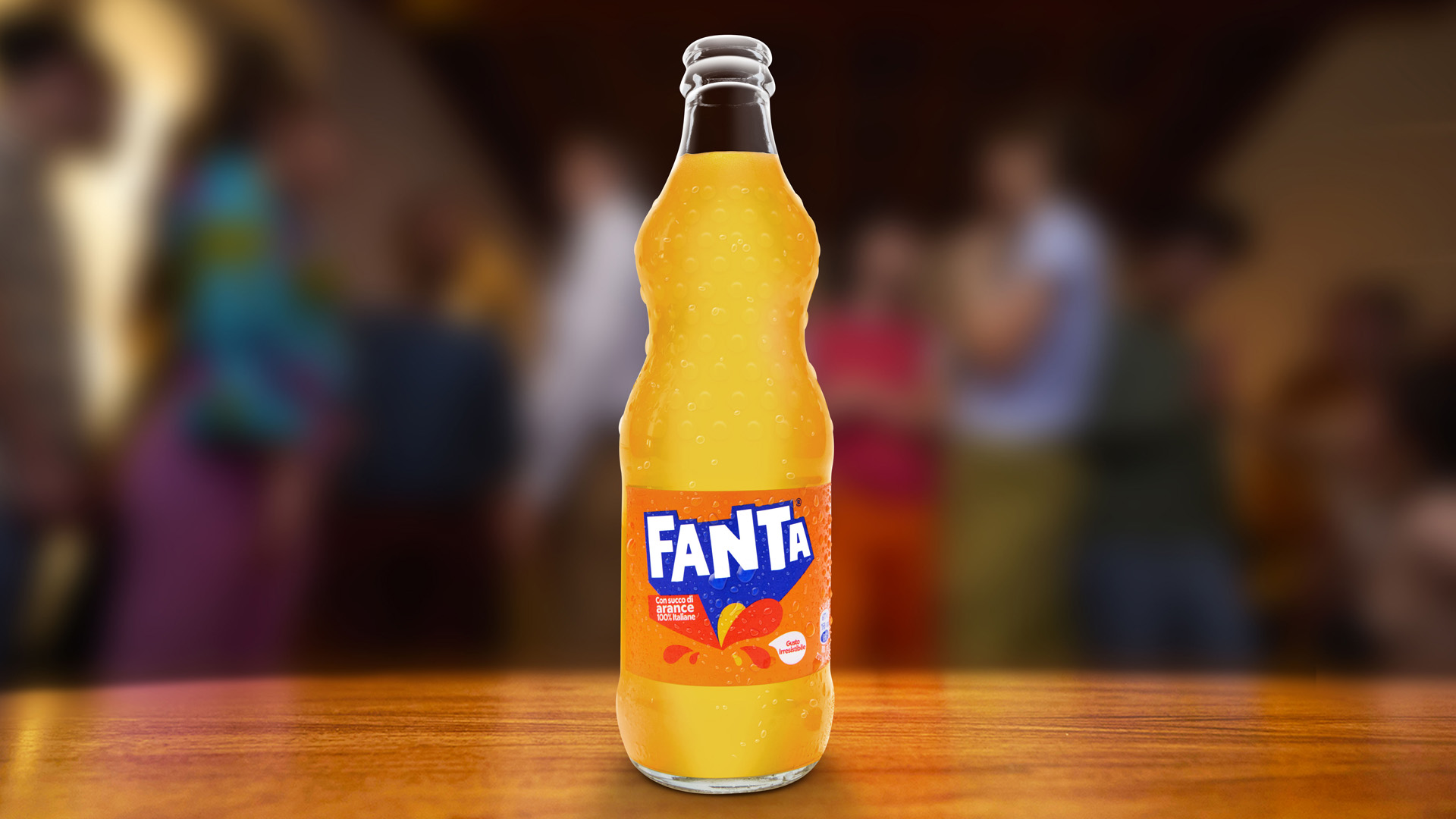 Bottiglia di Fanta con etichetta arancione e logo iconico, posizionata su un tavolo di legno. Sullo sfondo sfocato, un gruppo di persone in un’atmosfera conviviale.
