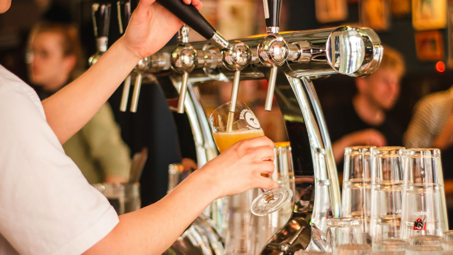 Un bartender serve una birra chiara alla spina in un bicchiere, mentre sullo sfondo si intravedono clienti seduti al bancone di un pub.
