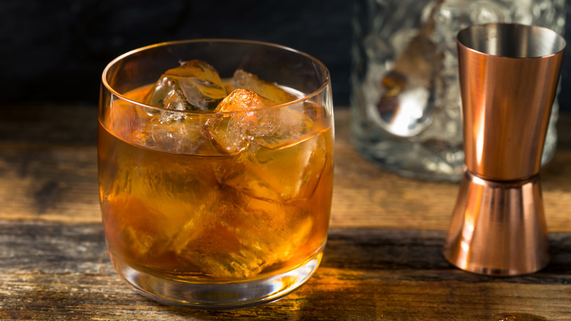 Cocktail French Connection servito in un bicchiere basso con ghiaccio, accanto a un jigger in rame e una bottiglia sfocata sullo sfondo, su un tavolo in legno rustico. Una ricetta cocktail a base di Cognac