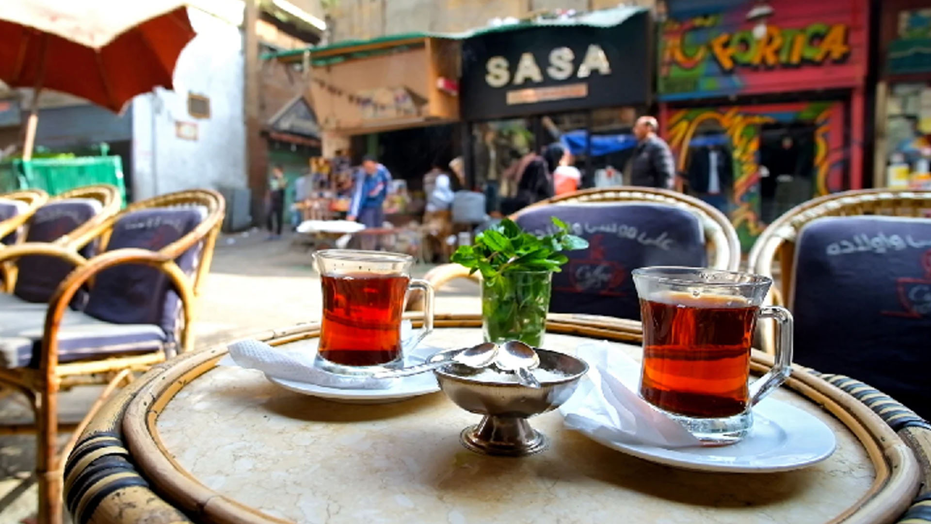 Due bicchieri di tè egiziano serviti su un tavolino in un tradizionale caffè all'aperto del Cairo. Sul tavolo, una ciotola con zollette di zucchero e un bicchiere di menta fresca. Sullo sfondo, sedie in vimini e insegne colorate animano la scena di un vivace mercato locale. Le bevande tradizionali del mondo Egitto
