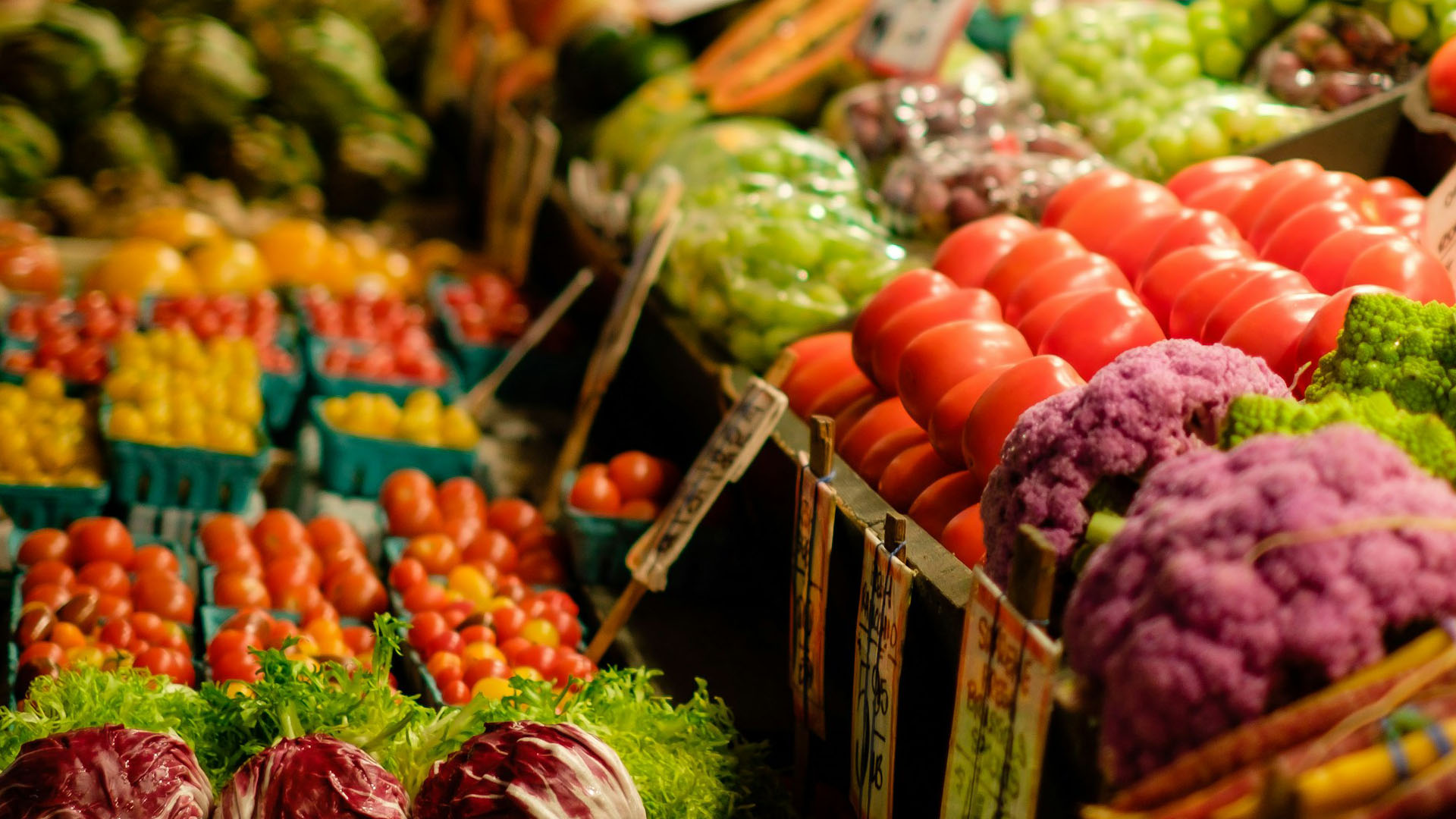 Un colorato mercato di prodotti freschi con una varietà di ortaggi e frutta in esposizione. In primo piano, pomodori rossi lucidi, cavolfiori viola e verdi, radicchio e lattuga. Sullo sfondo, cestini pieni di pomodorini gialli e rossi, oltre a verdure assortite, creando un'atmosfera vibrante e invitante tipica di un mercato agricolo o di una bottega di prodotti freschi. Tutti i Prodotti Agroalimentari Tradizionali italiani