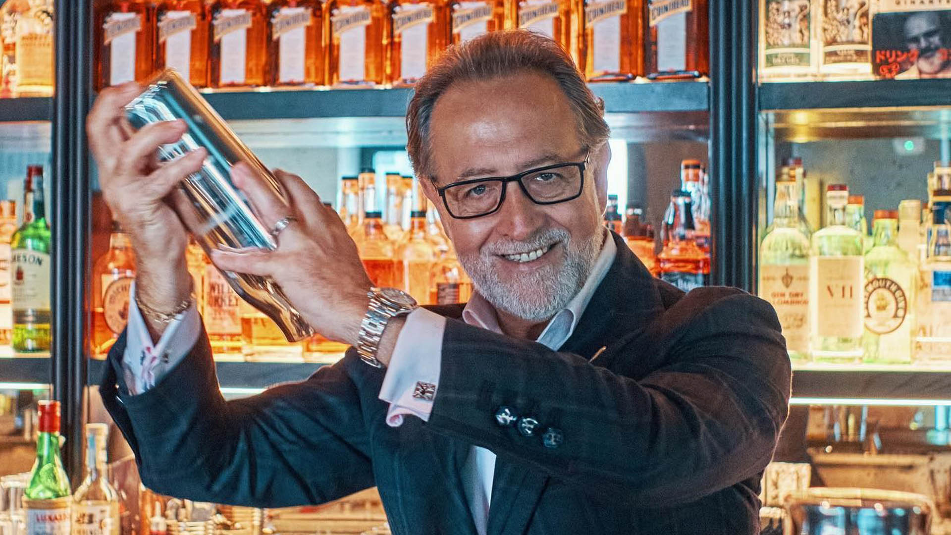 Salvatore Calabrese, celebre mixologist, sorride mentre agita uno shaker in un elegante bar, circondato da bottiglie di liquori pregiati. L’atmosfera trasmette passione e maestria nell’arte della mixology.
