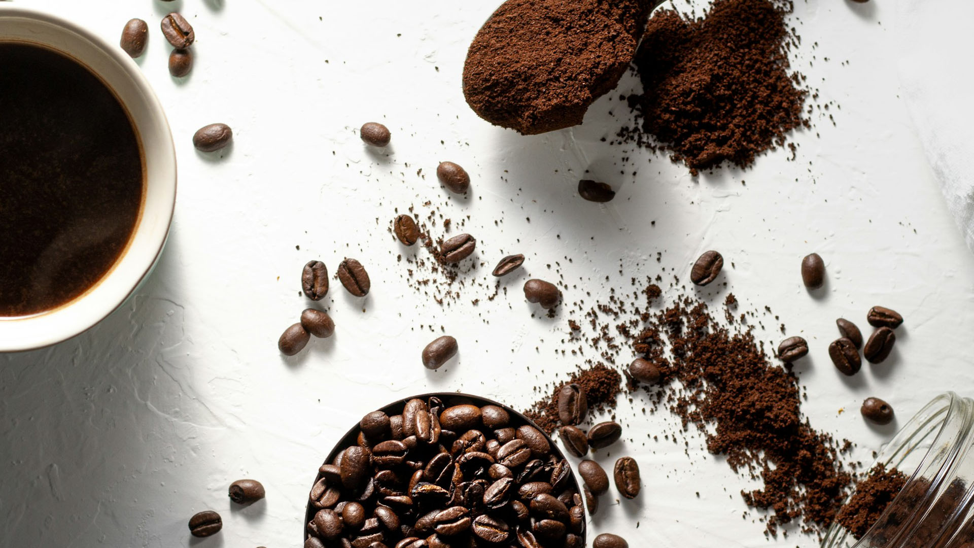 Chicchi di caffè interi e caffè macinato sparsi su una superficie bianca, accanto a una tazza di caffè pronto. Un'immagine che esalta la scelta tra caffè in grani o macinato, evidenziando la freschezza e l'aroma di entrambe le opzioni.