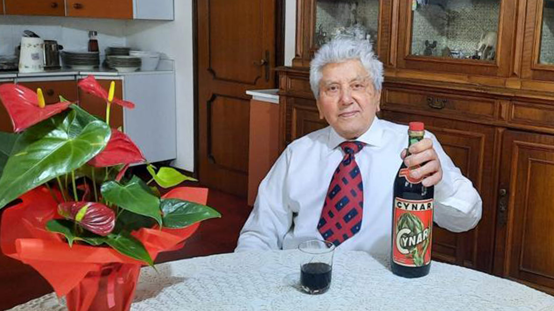 Rino Dondi Pinton, creatore del Cynar, seduto a un tavolo con una bottiglia dell'iconico liquore al carciofo e un bicchiere servito, in un ambiente domestico.