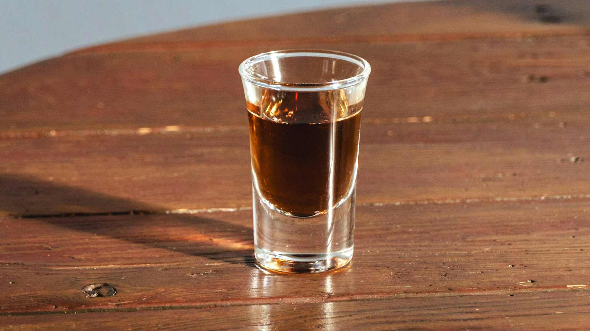 Bicchiere da shot riempito con un liquore scuro, posato su un tavolo in legno con luce calda che ne esalta i riflessi. Perfetto per degustazioni di amari. Guardando questa foto è impossibile capire le differenze tra amaro e liquore