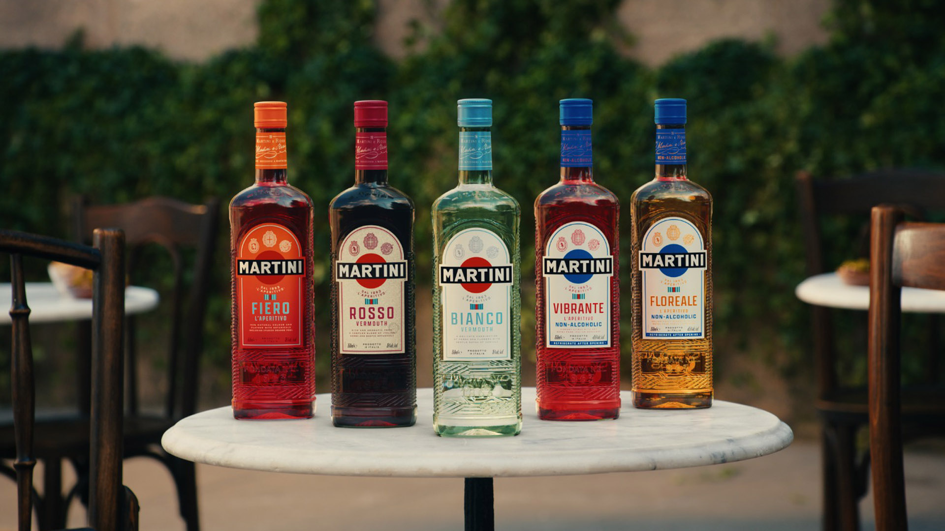 Cinque bottiglie della nuova gamma MARTINI posizionate su un tavolo all’aperto. Da sinistra a destra: MARTINI Fiero, MARTINI Rosso, MARTINI Bianco, MARTINI Vibrante (analcolico) e MARTINI Floreale (analcolico). Sullo sfondo, sedie e tavolini in un ambiente dall’atmosfera rilassata e conviviale