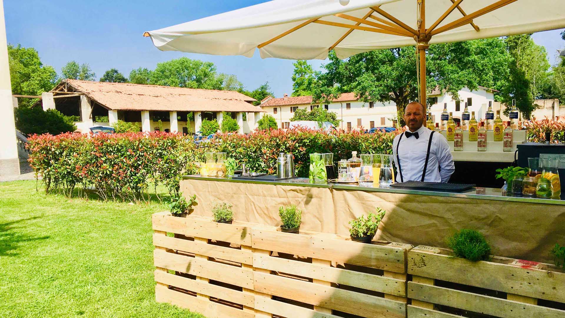 Bancone bar in stile rustico allestito per un evento all’aperto, con dettagli in legno e piante aromatiche. Barman elegante pronto a servire cocktail artigianali sotto un grande ombrellone bianco, in una location immersa nel verde. Barmanonline offre una soluzione di noleggio banconi per eventi.