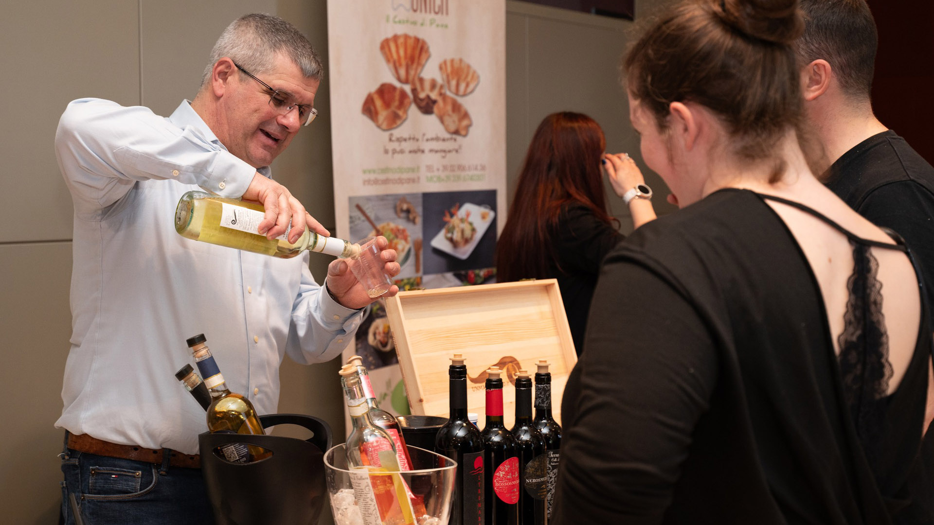 Un uomo in camicia chiara versa vino bianco in un bicchiere durante una degustazione a Ristoamare - Food & Drink Show 2025. Davanti a lui, alcune persone osservano e interagiscono. Sul tavolo, diverse bottiglie di vino e liquori, mentre sullo sfondo è visibile un banner con immagini di piatti gourmet