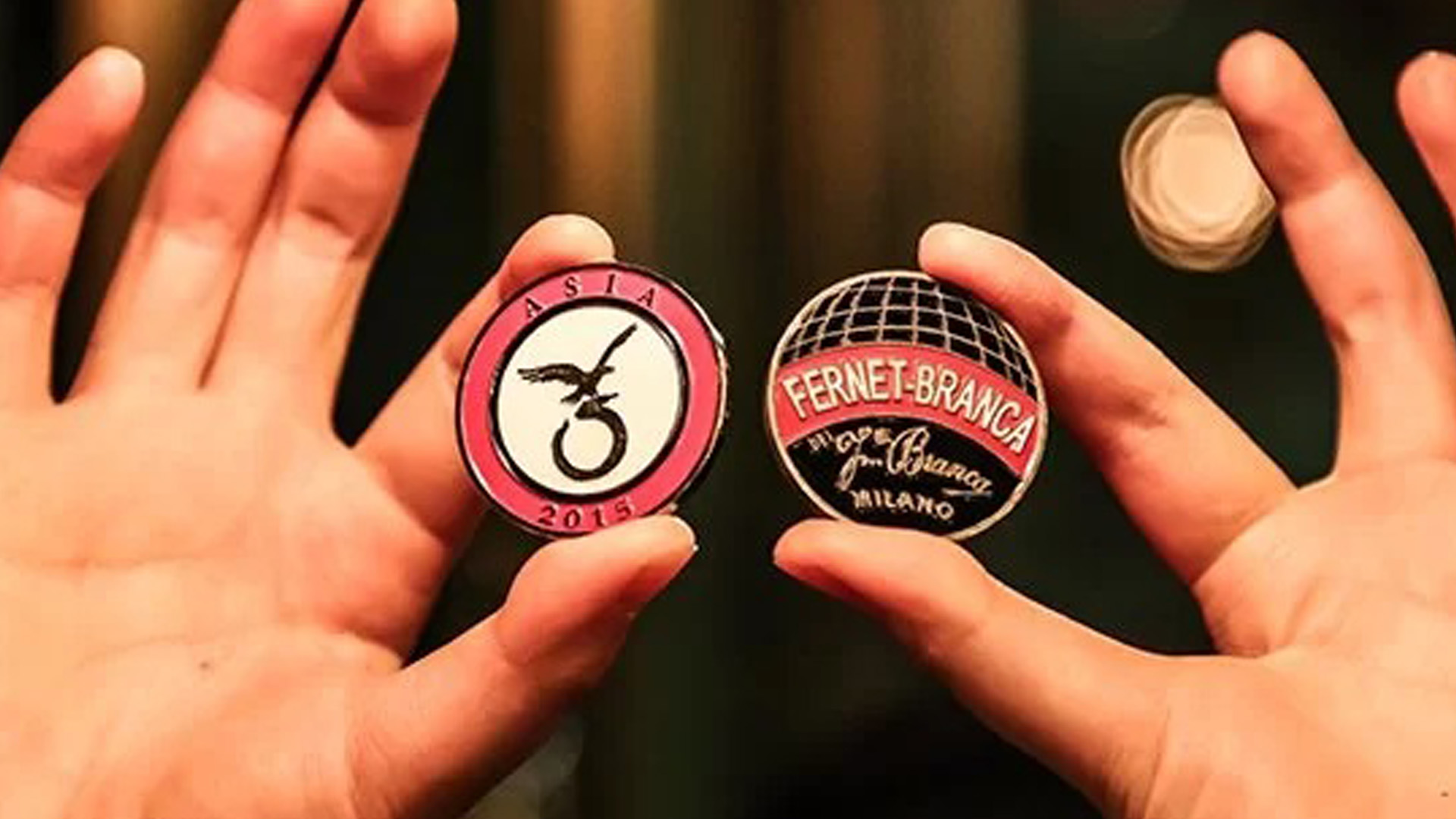 Due mani mostrano due Fernet Branca Coins, una dedicata all’Asia 2015 e l’altra con il logo storico Fernet Branca Milano, simboli esclusivi della community internazionale dei bartender.