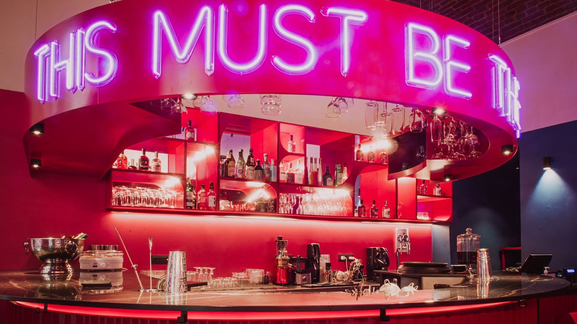 Interno del Formula n 12 Cocktail Bar a Torino con design moderno, arredamento rosso acceso e insegna al neon viola "This Must Be The Place" sopra al bancone.