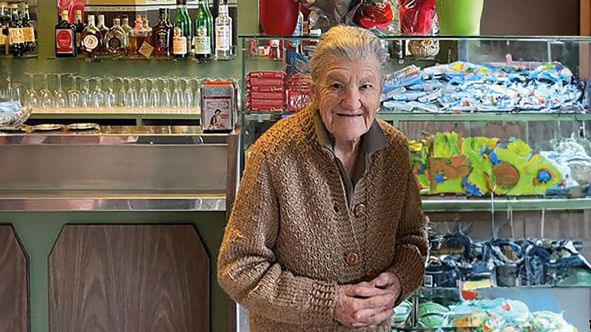 Anna Possi, la barista più anziana d'Italia, sorridente dietro il bancone del suo storico bar a Nebbiuno, con dolci artigianali in vetrina e bottiglie sugli scaffali.