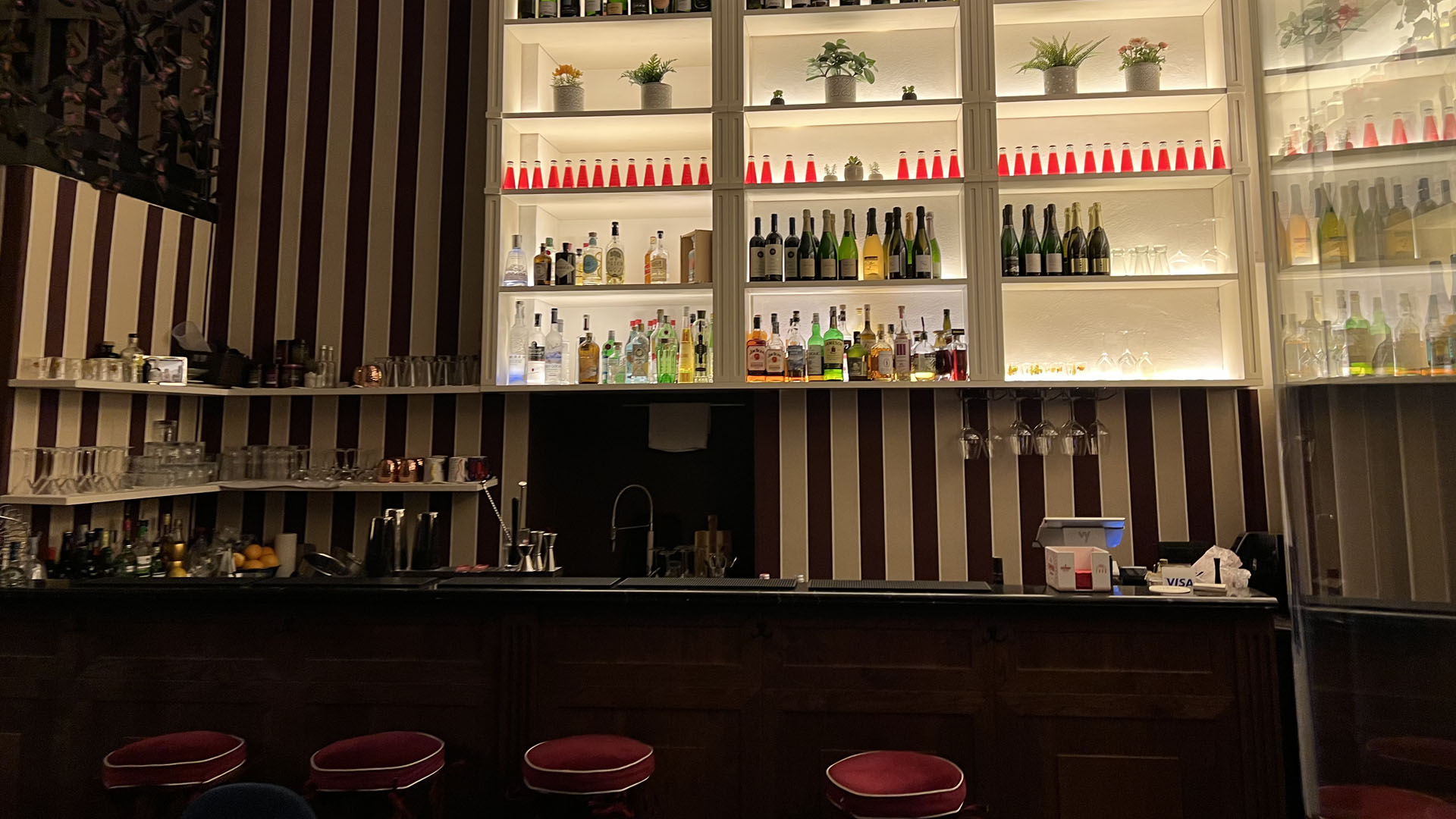 Interno elegante del Tipsy Cocktail Bar di Milano con bancone in legno scuro, sgabelli con seduta rossa, pareti a righe verticali e bottiglie esposte su mensole illuminate.