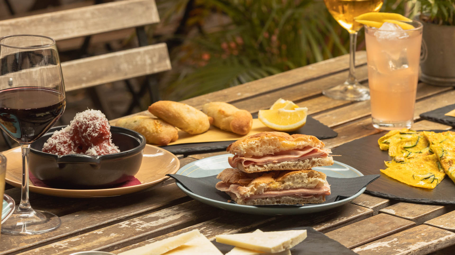 Tavolo all’aperto con specialità del Brunch romano di Städlin: pizza e mortazza, polpette al sugo con pecorino, frittata di zucchine, formaggi, pane caldo e cocktail, serviti in una location rustica e accogliente.
