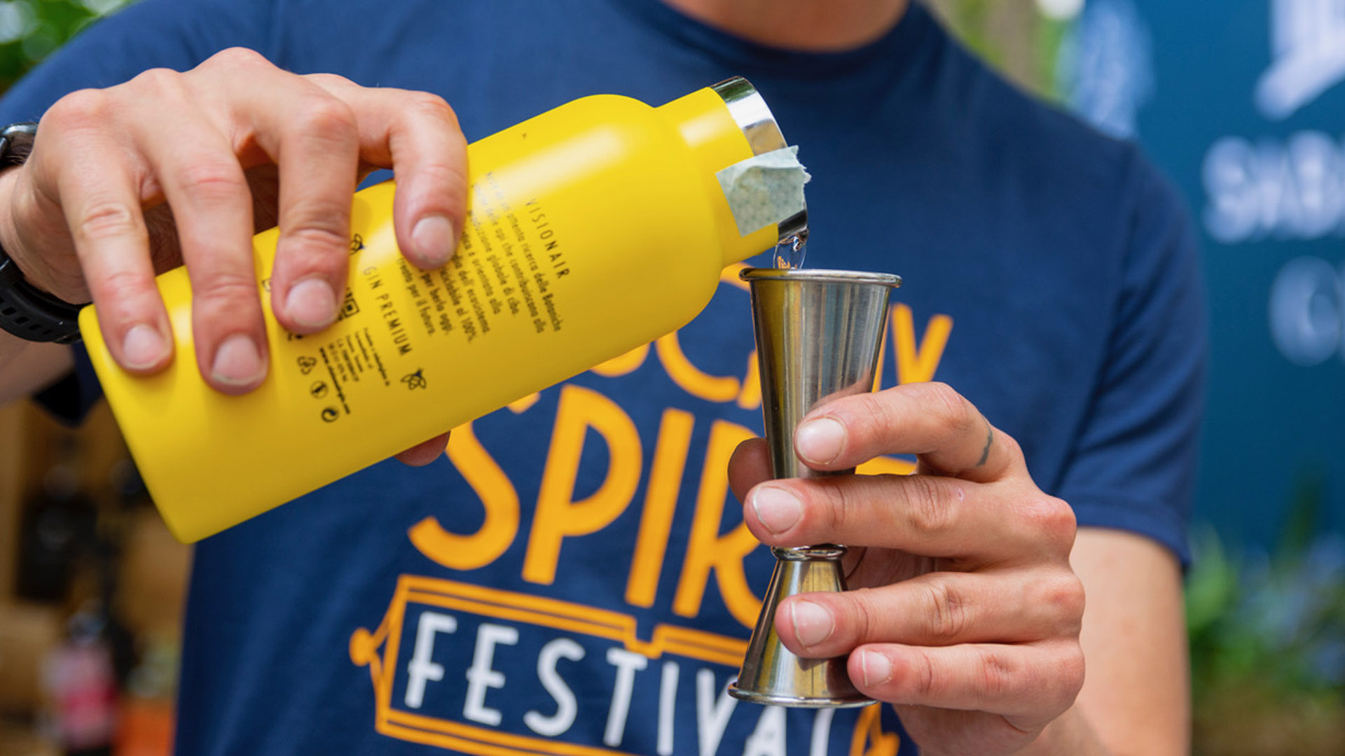 Primo piano di un bartender che versa gin da una bottiglia gialla in un jigger durante il Tuscany Spirit Festival 2025, con maglietta blu ufficiale dell’evento visibile sullo sfondo.