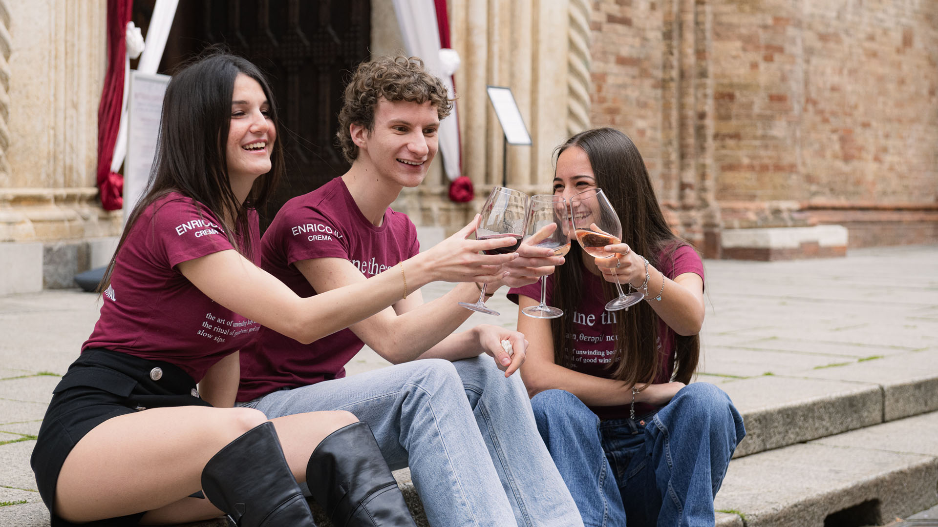 Tre giovani brindano sorridenti con calici di vino durante il Crema Wine Festival 2025, seduti all'aperto davanti a una chiesa storica. Indossano magliette coordinare dell’evento.