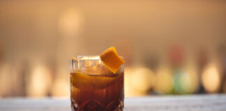 Aperitivo e caffè: quando l’espresso entra nella mixology Aperitivo e caffè nella mixology con cocktail scuro, scorza d’arancia e chicchi di caffè su bancone in legno.