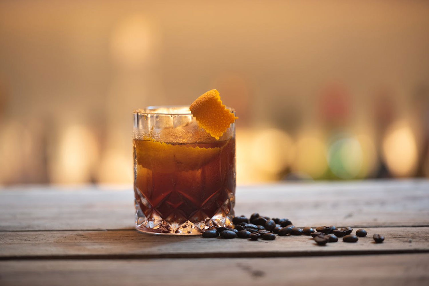 Aperitivo e caffè nella mixology con cocktail scuro, scorza d’arancia e chicchi di caffè su bancone in legno.