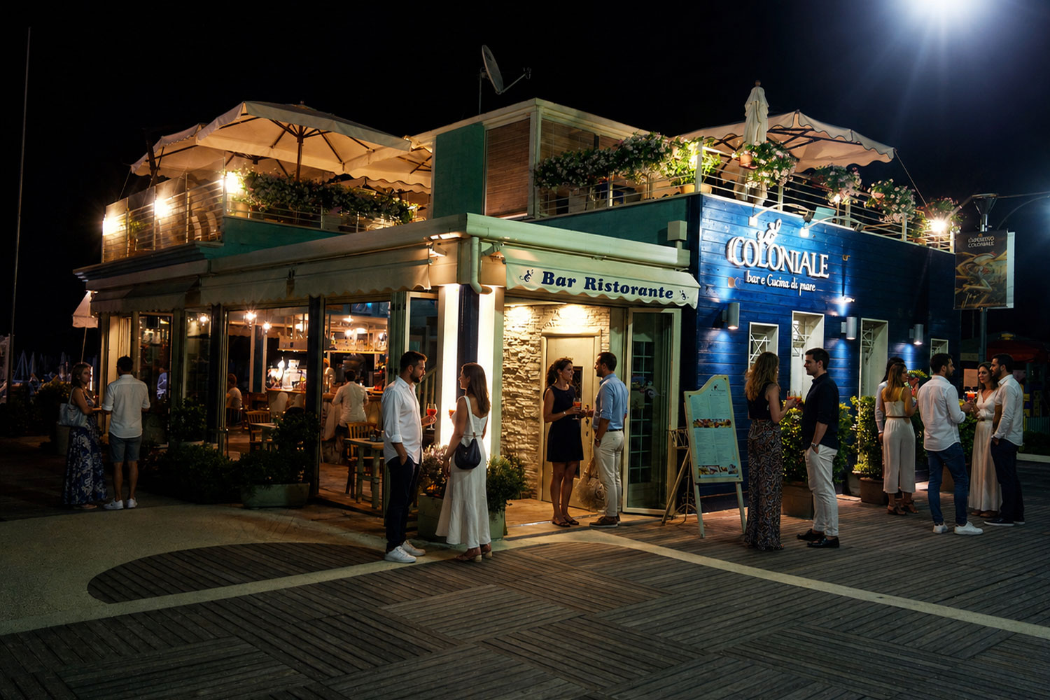 Il Coloniale a Cattolica di sera con ospiti in piedi e cocktail davanti al locale di Altamarea Beach Village