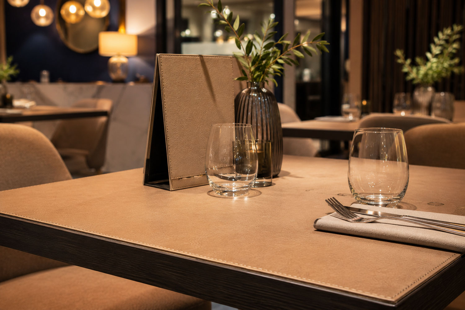 Cover per tavolo su misura in ecopelle con bordo cucito e mise en place elegante all’interno di un moderno ristorante, bar o hotel.